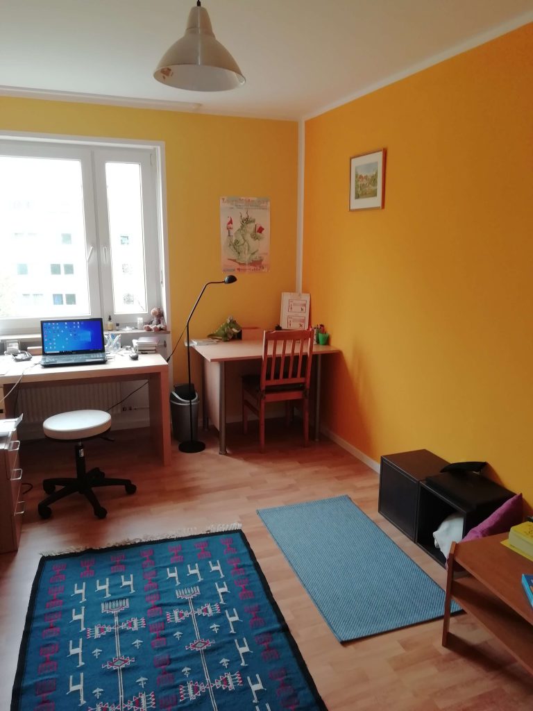 Hier sehen Sie den Praxisraum von LRS Wortschatz. Es befinden sich dort zwei Schreibtische mit Lernmaterialien und einem Laptop. Die Wände sind in einem warmen Orange gestrichen und mit Bildern dekoriert.